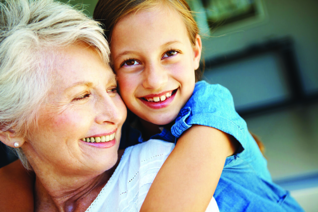 a young girl smiles happily while hugging her grandmother from behind